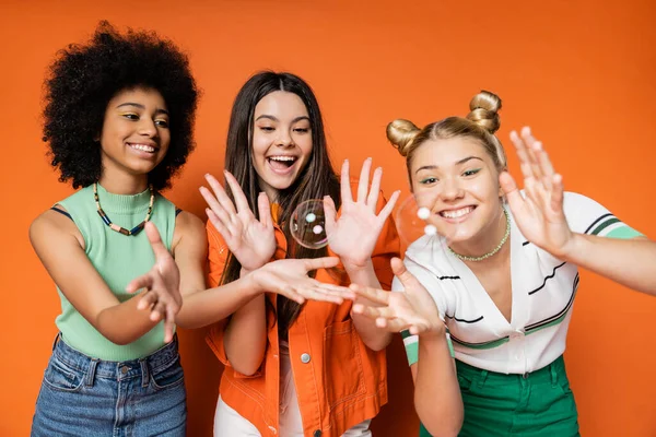 stock image Cheerful and multiethnic teen girlfriends in stylish casual clothes looking at soap bubbles while standing on orange background, teen fashionistas with impeccable style concept