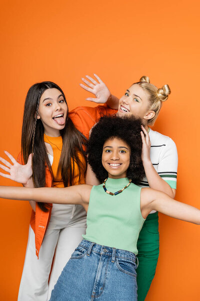 Cheerful and multiethnic teenage girlfriends with colorful makeup posing in casual outfits and looking at camera together while standing isolated on orange, fashionable and trendy clothes