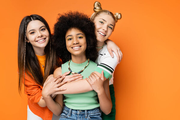 Positive girlfriends with colorful makeup hugging stylish african american girlfriend and looking at camera together while standing isolated on orange, fashionable and trendy clothes