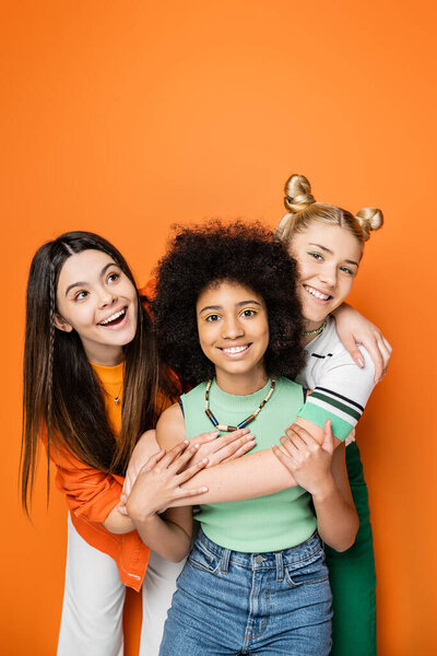 Positive teenagers with colorful makeup and casual outfits hugging african american girlfriend together and posing on orange background, fashionable and trendy clothes