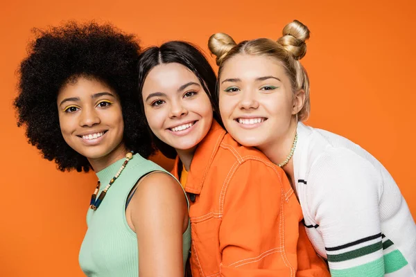 stock image Portrait of cheerful and multiethnic stylish teenage girlfriends with bold makeup wearing casual clothes and looking at camera together on orange background, stylish and confident poses