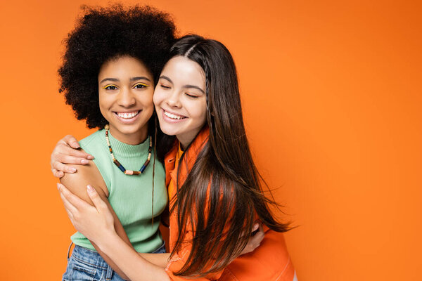 Cheerful brunette teenage girl in casual outfit hugging trendy african american girlfriend with colorful makeup and standing together on orange background, stylish and confident poses