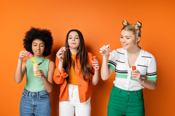 Cheerful blonde teenage girl in casual clothes holding soap bubbles near multiethnic girlfriends with colorful makeup while standing on orange background, trendy and stylish hairstyles