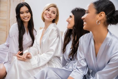 bridal party, silk robes, female friends, bride with her multicultural bridesmaids, brunette and blonde women, cultural diversity, happy girlfriends sitting on bed, positivity, wedding preparations  clipart