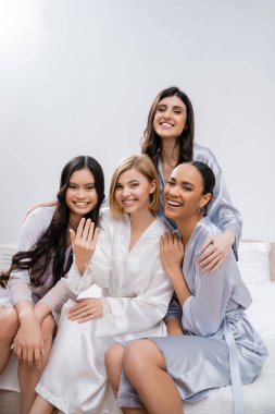 Dört kadın, gelin partisi, mutlu sarışın gelin ırklar arası nedimelerinin yanında nişan yüzüğü gösteriyor, yatakta oturuyor, mutluluk, ipek bornoz, esmer ve sarışın, en iyi arkadaşlar, çeşitlilik.
