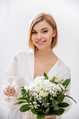 Beyaz ipek bornozlu neşeli gelin gülümsüyor, elinde şampanya kadehi ve gelin buketi, genç kadın, güzel, heyecanlı, kadınsı, mutlu, portre 