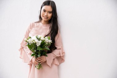Pastel pembe elbiseli neşeli nedime elinde buket, gri arka planda esmer Asyalı kadın, beyaz çiçekler, özel bir gün, düğün, moda, gülümseme ve neşe, kameraya bakıyor. 
