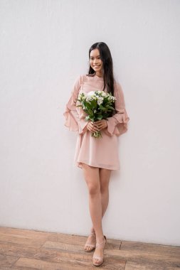 Pastel pembe nedime elbiseli mutlu Asyalı kadın elinde buket, esmer saçlı, gri arka planda poz veren, beyaz çiçekler, özel bir gün, düğün, moda, gülümseme ve neşe, tam boy 