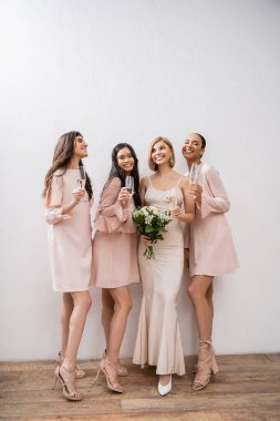 Joy, gelinlik giymiş sarışın gelin elinde buket, ırklar arası nedimeler, şampanya kadehleri, gri arka plan, ırksal çeşitlilik, moda, çok kültürlü genç kadınlarla duruyor. 