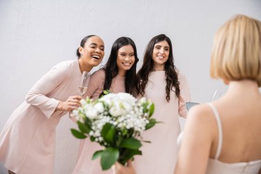 wedding preparations, cheerful multicultural bridesmaids with champagne looking at blonde bride on grey background, admire her style, fitting, bridesmaid gowns, diversity, blurred, special occasion  clipart