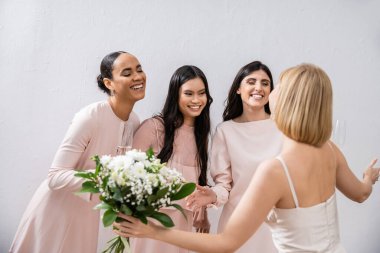 wedding preparations, cheerful multicultural bridesmaids with champagne looking at blonde bride with bouquet on grey background, admire her style, fitting, bridesmaid gowns, diversity  clipart