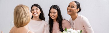wedding preparations, cheerful interracial bridesmaids looking at blonde bride with bouquet on grey background, admire her style, fitting, bridesmaid gowns, diversity, banner  clipart