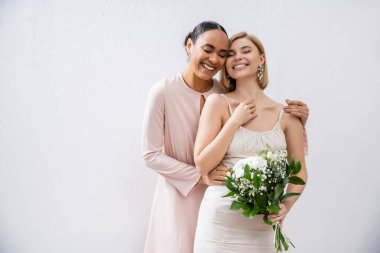 special occasion, happy bride with bridesmaid, positive interracial women, wedding dress and bridesmaid gown, african american woman hugging engaged friend on grey background  clipart