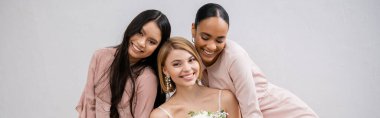 wedding photography, cultural diversity, three women, happy bride with bouquet and her interracial bridesmaids sitting on armchair on grey background, brunette and blonde, joy, celebration, banner  clipart