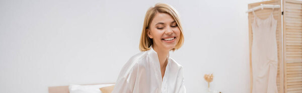 cheerful bride with blonde hair sitting in white silk robe on bed, smiling, wedding planning, young woman, beautiful, excitement, feminine, blissful, preparation, pre-wedding, banner 