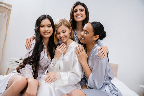 four women, bridal party, joyful blonde bride and her interracial bridesmaids sitting on bed together, happiness, silk robes, engagement ring, brunette and blonde, best friends, diversity, laughter 