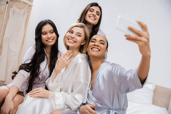 четыре женщины, свадебная вечеринка, веселая блондинка невеста и ее межрасовые подружки невесты делают селфи вместе, счастье, шелковые одежды, обручальное кольцо, брюнетка и блондинка, лучшие друзья, разнообразие