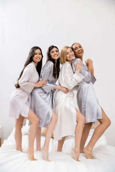 stock image bridal shower, four women, cheerful bride and bridesmaids in silk robes standing on bed, cultural diversity, having fun together, friendship goals, brunette and blonde women, looking at camera 