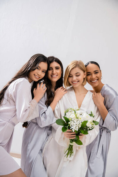 bridal party, four women, joyful bride holding bouquet with white flowers near bridesmaids in silk robes, cultural diversity, togetherness, friendship goals, brunette and blonde women 