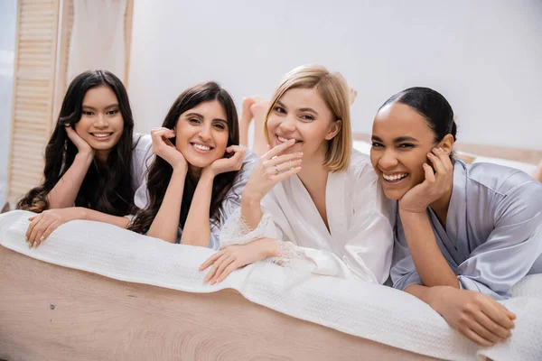 stock image positivity, bridal party, four women, happy bride and bridesmaids in silk robes lying on bed, looking at camera, cultural diversity, having fun together, friendship goals, brunette and blonde women 