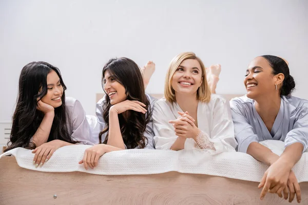 stock image happiness, bridal party, four women, happy bride and bridesmaids in silk robes lying on bed, looking away, cultural diversity, having fun together, friendship goals, brunette and blonde women 