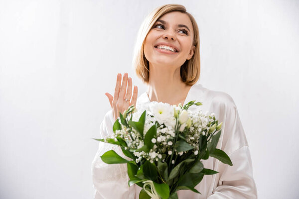 white flowers, happiness, cheerful bride with blonde hair standing in white silk robe and holding bridal bouquet, showing engagement ring, young woman, beautiful, excitement, feminine, blissful 