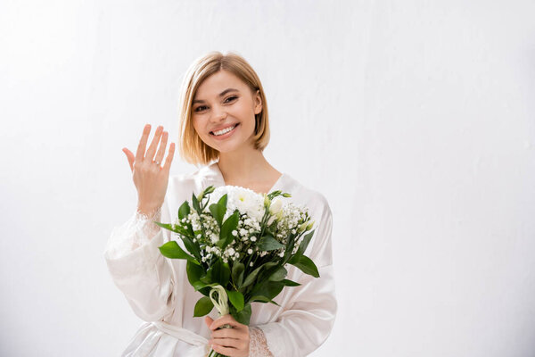 happiness, cheerful bride with blonde hair standing in white silk robe and holding bridal bouquet, showing engagement ring, young woman, beautiful, excitement, feminine, blissful, white flowers 