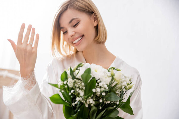 happiness, cheerful bride with blonde hair wearing white silk robe and holding bridal bouquet, showing engagement ring, young woman, beautiful, excitement, feminine, blissful, white flowers 