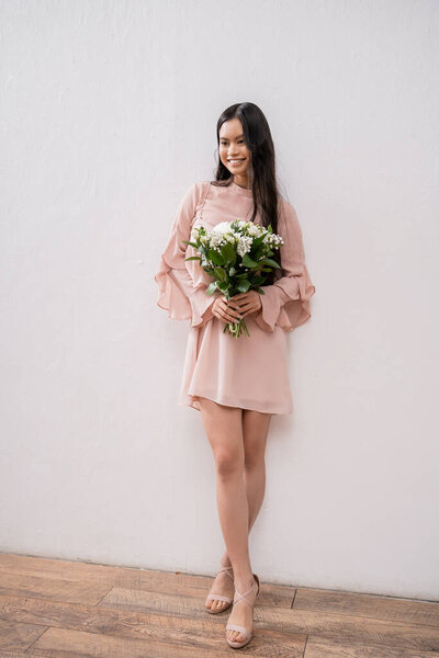 счастливая азиатка в пастельно-розовом платье подружки невесты держа букет, брюнетки волосы, позируя на сером фоне, белые цветы, особый случай, свадьба, мода, улыбка и радость, полная длина 