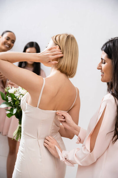 подготовка к свадьбе, счастливая подружка невесты застегивая молнию свадебное платье блондинки невесты, межрасовые женщины на размытом сером фоне, расовое разнообразие, мода, брюнетки и блондинки женщины, свадебный букет 