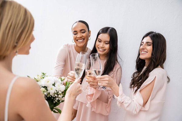 happy interracial girlfriends clinking glasses with champagne, bride with white flowers, brunette and blonde women, bridesmaids, diversity, positivity, bridal bouquet, grey background 
