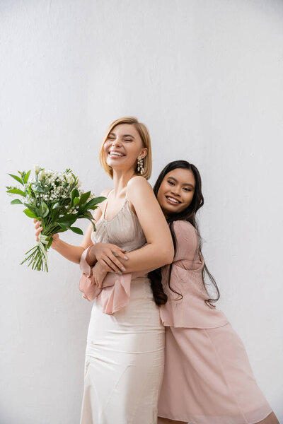 особый случай, азиатская подружка невесты обнимая красивую невесту, цели дружбы, серый фон, счастливые подруги, свадебный букет, блондинка и брюнетка женщины, белые цветы, счастье 