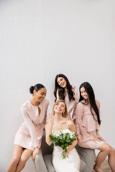wedding photography, diversity, four women, joyful bride with bouquet showing her engagement ring near bridesmaids, wedding day, sitting on armchair, grey background, happiness and joy 