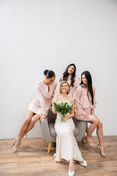 wedding photography, cultural diversity, four women, joyful bride with bouquet showing her engagement ring near bridesmaids, wedding day, sitting on armchair, grey background, happiness and joy 