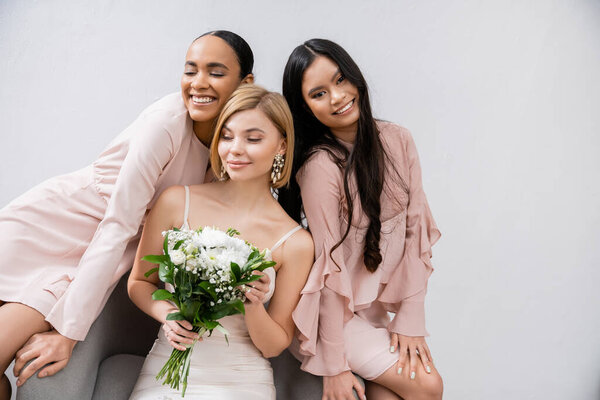 wedding theme, cultural diversity, three women, cheerful bride with bouquet and her interracial bridesmaids sitting on armchair on grey background, brunette and blonde, joy, celebration 