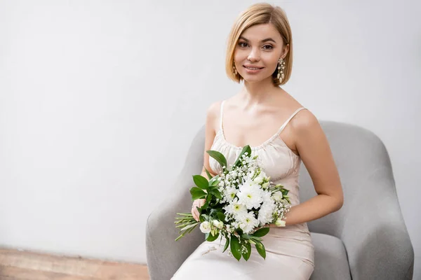 特別な機会に アームチェアに座って灰色の背景に花束を持っているウェディングドレスの美しいブロンドの花嫁 カメラ 白い花 ブライダルアクセサリー 女性を見て — ストック写真