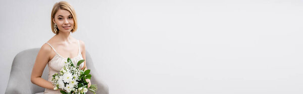 special occasion, beautiful, blonde bride in wedding dress sitting in armchair and holding bouquet on grey background, white flowers, bridal accessories, happiness, banner