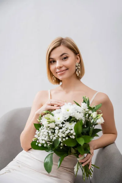 特別な機会に アームチェアに座って灰色の背景に花束を持っているウェディングドレスの美しいブロンドの花嫁 婚約指輪 白い花 ブライダルアクセサリー — ストック写真