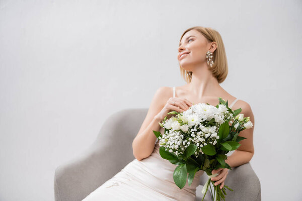 special occasion, happy blonde bride in wedding dress sitting in armchair and holding bouquet on grey background, engagement ring, white flowers, bridal accessories, happiness, feminine 