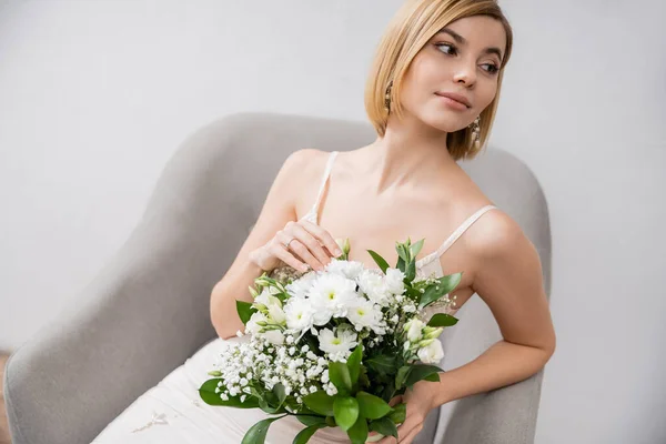 特別な機会に アームチェアに座って灰色の背景 婚約指輪 白い花 ブライダルアクセサリー 女性の花束を保持するウェディングドレスの美しい若い花嫁 — ストック写真