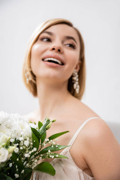 special occasion, beautiful, blonde bride in wedding dress holding bouquet on grey background, white flowers, bridal accessories, happiness, feminine, blissful, looking away, joy 