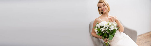 cheerful and blonde bride in wedding dress sitting in armchair and holding bouquet on grey background, white flowers, bridal accessories, happiness, special occasion, beautiful, feminine, banner