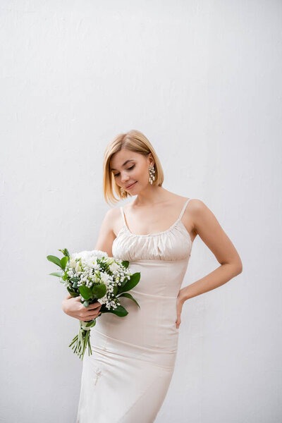 happy and blonde bride in wedding dress holding bouquet on grey background, white flowers, bridal accessories, happiness, special occasion,   beautiful, feminine, blissful 