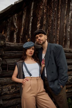 Stylish bearded man in jacket and Newsboy Cap looking at camera and posing while standing near girlfriend in vintage outfit and suspenders and countryside house, couple in love enjoying nature clipart