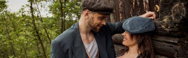 Smiling and fashionable man in jacket and newsboy cap looking at cheerful girlfriend while standing together near rustic house at nature, stylish couple in rural setting, banner  clipart