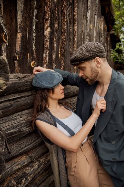 Fashionable and passionate woman in suspenders and newsboy cap touching jacket of bearded boyfriend while standing together near rustic house, stylish couple in rural setting clipart