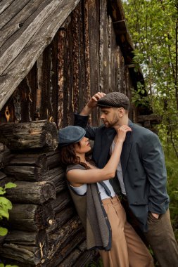 Fashionable woman in vintage vest and suspenders touching shoulder of bearded boyfriend in jacket and newsboy cap while standing near rural house, stylish couple in rural setting clipart