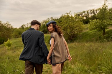 Fashionable brunette woman in vest and newsboy cap looking at camera and holding hand of bearded boyfriend and walking with landscape at background, stylish couple in rural setting clipart