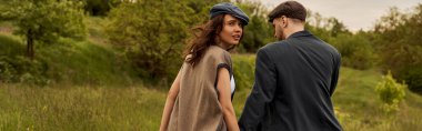 Trendy brunette woman in vest and newsboy cap looking at camera while walking near boyfriend in jacket with blurred landscape at background, stylish couple in rural setting, banner  clipart