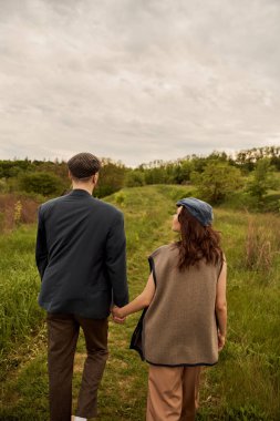 Brunette and fashionable woman in vest and newsboy cap holding hand of boyfriend in jacket while walking together on meadow with nature and sky at background, fashion-forwards in countryside clipart